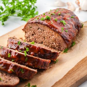 Side view of air fryer meatloaf sitting on a wood cutting board sliced like a loaf of bread. A red ketchup based glaze on top and garnished with parsley.
