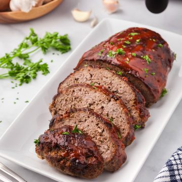 Side shot of air fryer meatloaf, half of which have been cut into slices. The top has a red BBQ glaze and is garnished with parsley flakes. The air fryer is peeking in in the upper right corner.