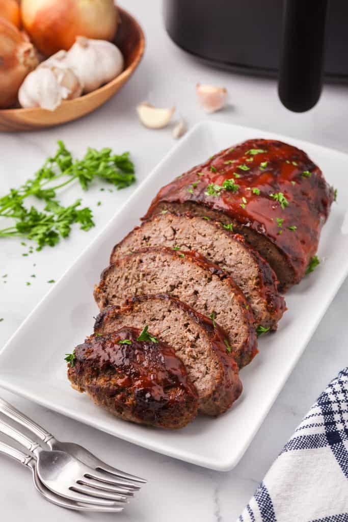 Side shot of air fryer meatloaf, half of which have been cut into slices. The top has a red BBQ glaze and is garnished with parsley flakes. The air fryer is peeking in in the upper right corner.