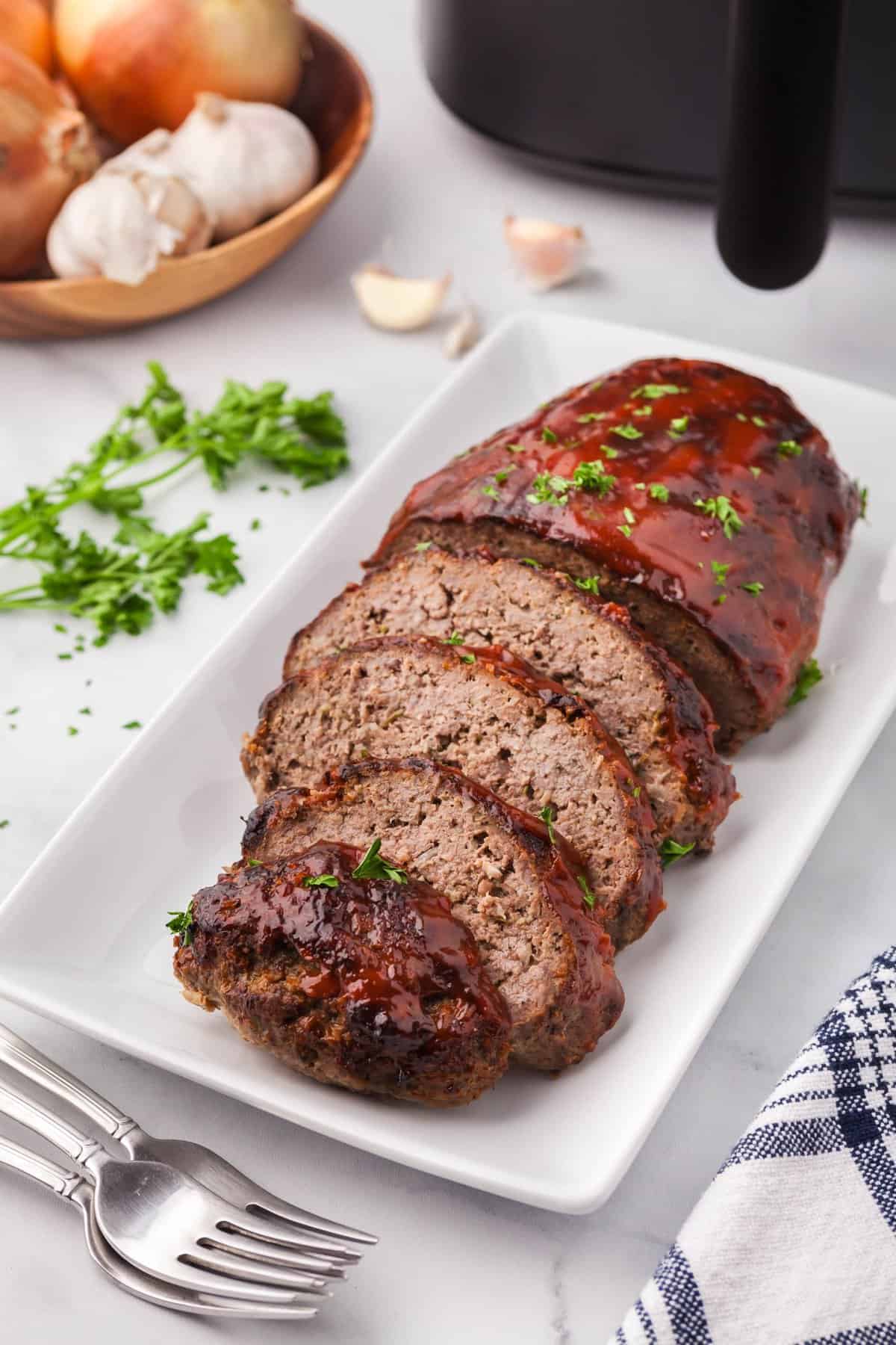 Side shot of air fryer meatloaf, half of which have been cut into slices. The top has a red BBQ glaze and is garnished with parsley flakes. The air fryer is peeking in in the upper right corner.