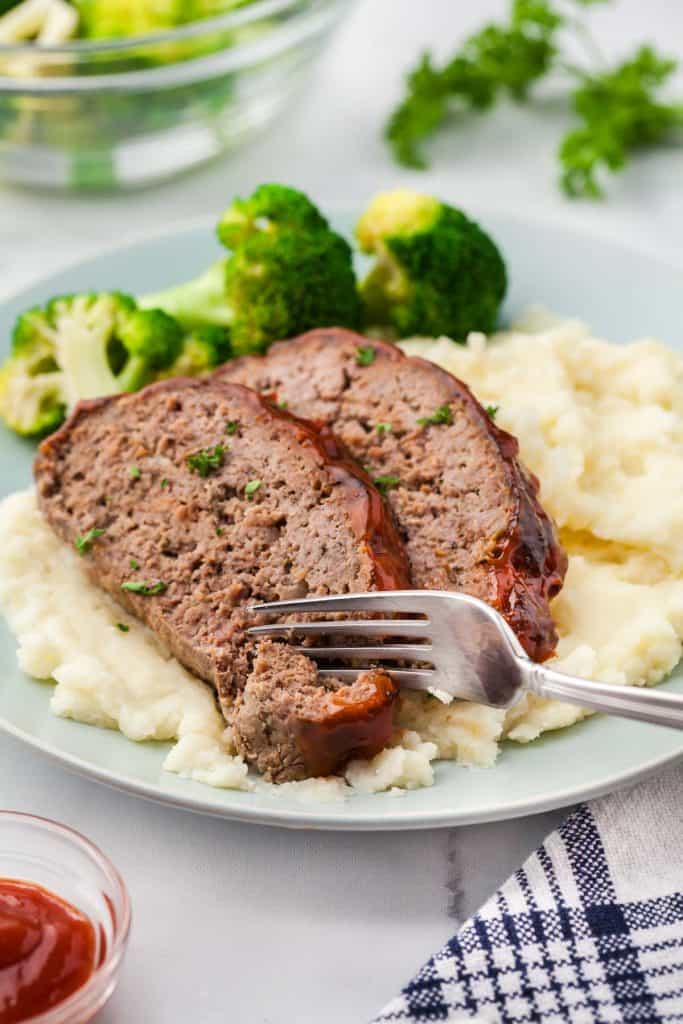 A fork is slicing into one of two slices of air fryer meatloaf laying atop of mashed potatoes with steamed broccoli on a pale blue plate. A small serving of glaze is in the foreground for dipping.