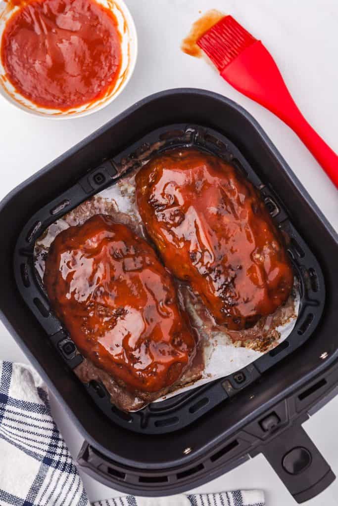 Air fryer basket containing two cooked meatloaves which have been coated in the ketchup based glaze and ready to be caramelized. Above the basket is a small bowl of glaze and a red silicone basting brush.