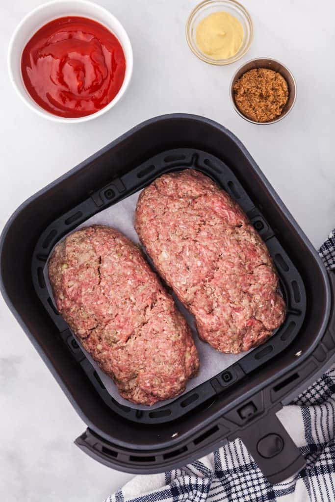 Air fryer basked lined with parchment and containing two shaped meatloaves ready to be cooked. Above is a small bowl of ketchup, a small metal container of brown sugar, and a glass pinch bowl of dijon mustard.