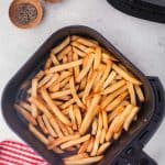 overhead shot of air fryer frozen french fries sitting in the air fryer basket with the air fryer in the top left hand corner, and wooden pinch bowls of salt and pepper in the top right hand corner.
