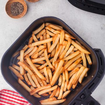 overhead shot of air fryer frozen french fries sitting in the air fryer basket with the air fryer in the top left hand corner, and wooden pinch bowls of salt and pepper in the top right hand corner.