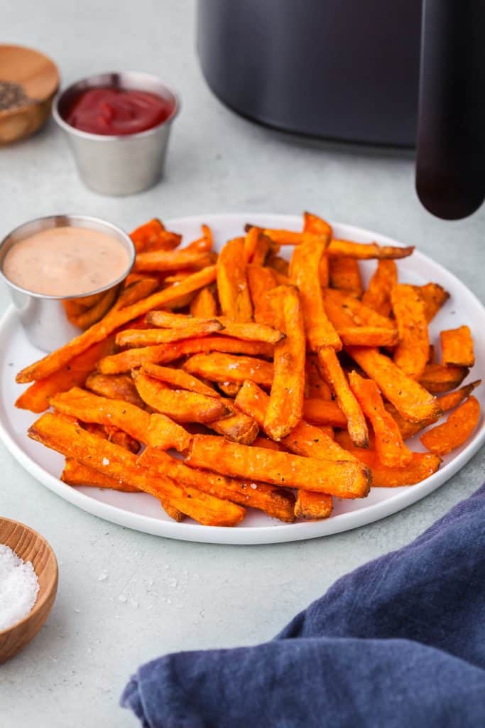 Sweet potato fries served on a white plate with a black air fryer in the background.  Ketchup and chipotle mayo served as dipping sauces