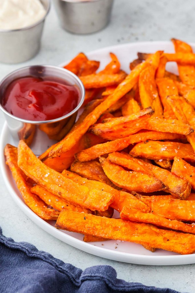 Side shot of a white ceramic plate filled with bright orange sweet potato fries. Nestled on the edge is a small stainless steel bowl of ketchup. Off the plate are two small stainless steel pinch bowls of mayonnaise and chipotle mayonnaise for dipping.