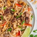 Close up overhead shot of beef pad thai served on a white plate. Crushed peanuts in a small bowl in the upper right corner and green onion pieces in the bottom right corner of the scene.