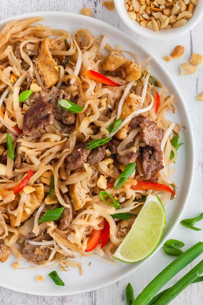 Close up overhead shot of beef pad thai served on a white plate. Crushed peanuts in a small bowl in the upper right corner and green onion pieces in the bottom right corner of the scene.