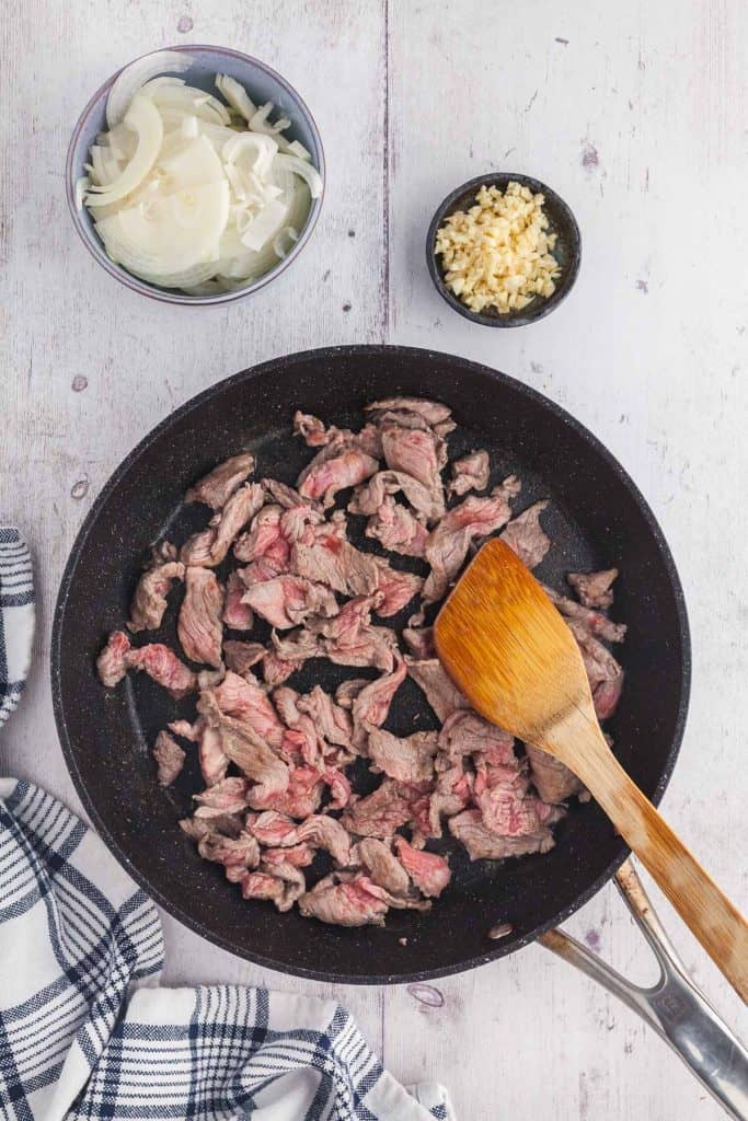 Overhead shot of a large fry pan stir frying thin strips of tender beef. Small bowls of garlic and onion above are waiting to be added to the pan