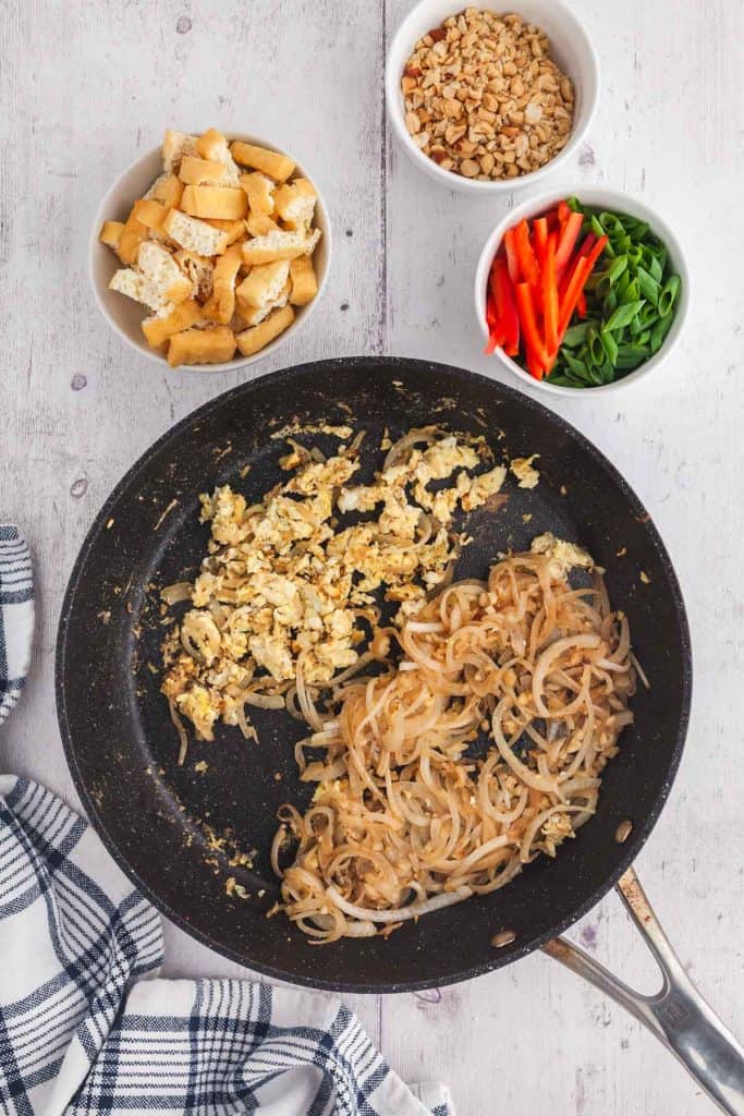 Overhead shot of large fry pan with cooked onions pushed to the bottom right side while the top left side was used to scramble an egg. The next ingredients to be added are in small bowls above, tofu, green onions, red peppers, and crushed peanuts.