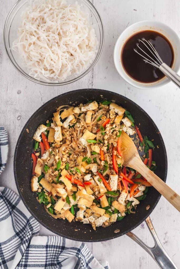 Overhead shot cooking beef pad thai in a large fry pan. All ingredients are in the pan except for the sauce and soft rice noodles which are in small bowls above the fry pan.