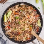 Overhead shot of Beef Pad Thai just finished cooking in a large fry pan with a wooden spatula ready to serve. White plates with chopsticks in the upper left corner and lime wedges for serving in the upper right corner.