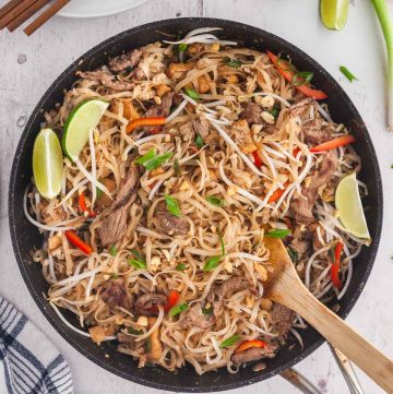 Overhead shot of Beef Pad Thai just finished cooking in a large fry pan with a wooden spatula ready to serve. White plates with chopsticks in the upper left corner and lime wedges for serving in the upper right corner.