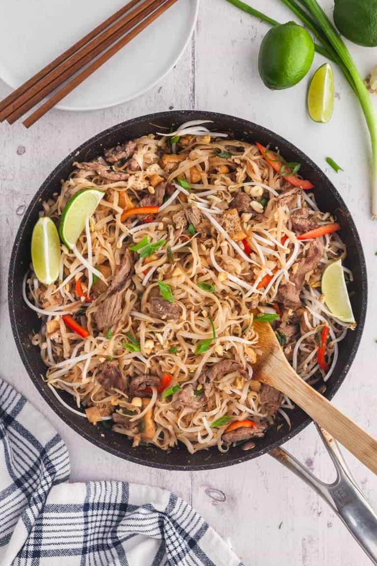 Overhead shot of Beef Pad Thai just finished cooking in a large fry pan with a wooden spatula ready to serve. White plates with chopsticks in the upper left corner and lime wedges for serving in the upper right corner.