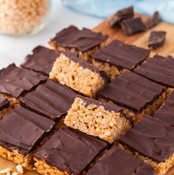 chocolate and peanut butter rice krispie treats cut into squares and placed on a wooden cutting board. Two of the inner squares are tilted up on their edge revealing the crispy rice krispies underneath a layer of chocolate