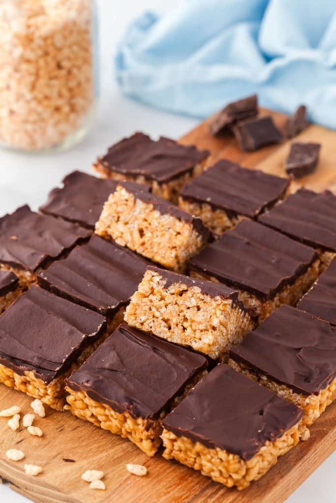 chocolate and peanut butter rice krispie treats cut into squares and placed on a wooden cutting board. Two of the inner squares are tilted up on their edge revealing the crispy rice krispies underneath a layer of chocolate