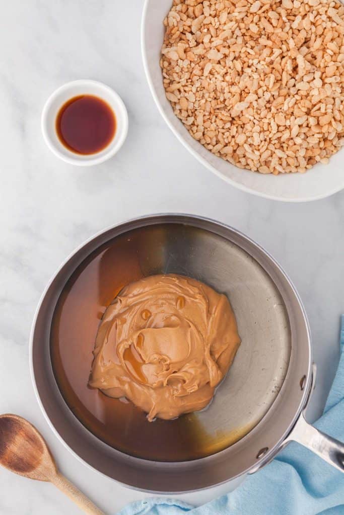An overhead shot of a small saucepan containing the peanut butter, maple syrup, and corn syrup. A pinch bowl of vanilla and a large bowl of rice krispies are above the saucepan, and a wooden spoon is poking into the bottom right corner of the shot.