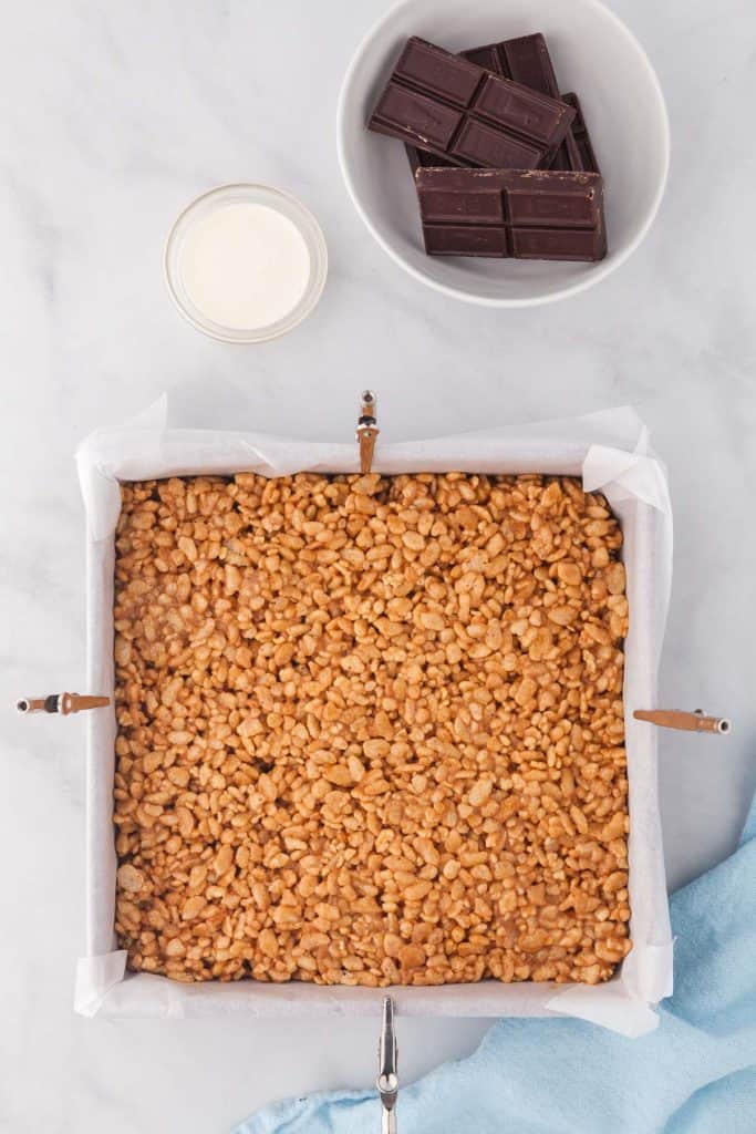 overhead shot of a parchment sheet lined 8x8 pan containing the homogenous peanut butter rice krispie mixture. Above the pan is the heavy cream and chocolate which will be used to make the chocolate ganache topping