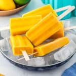 A blue shallow bowl filled with ice holding six mango popsicles. Background has a wooden bowl filled with mangos and limes.