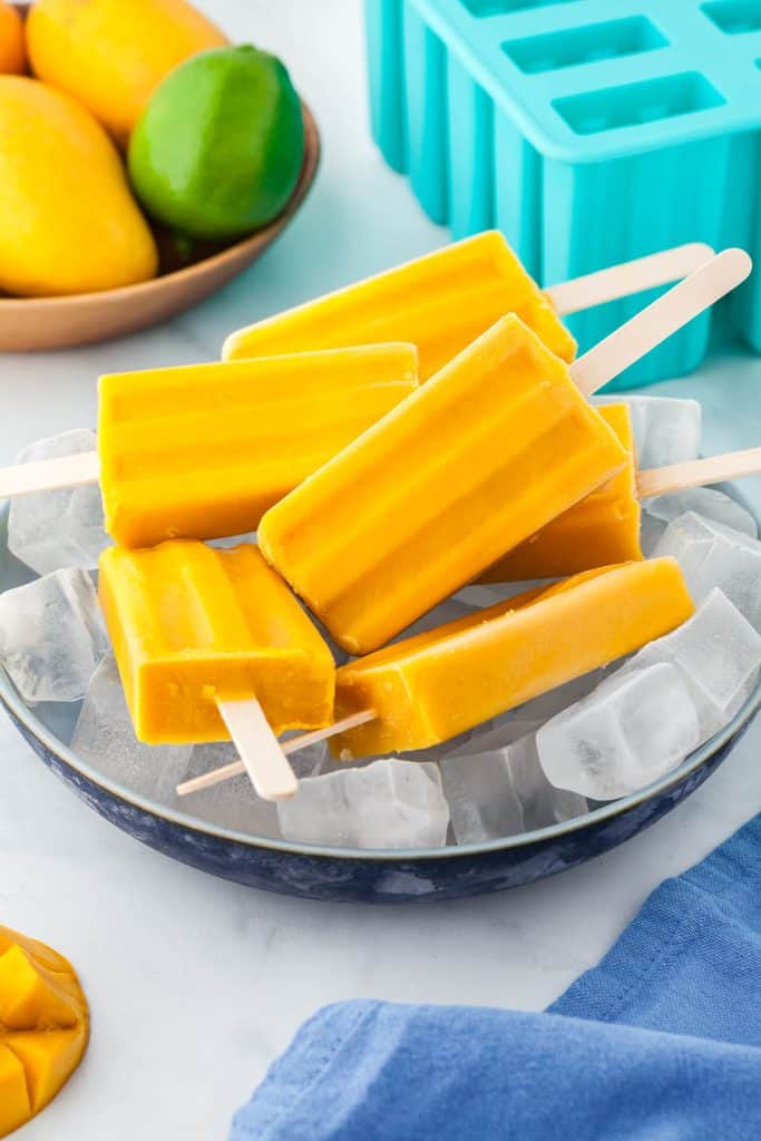 A blue shallow bowl filled with ice holding six mango popsicles.  Background has a wooden bowl filled with mangos and limes.