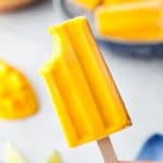 One mango popsicle with bite out of the top left corner being held up in the middle of the photo. In the background there is a shallow bowl filled with ice containing other mango popsicles, and there is a sliced fresh mango on the left side in the background.
