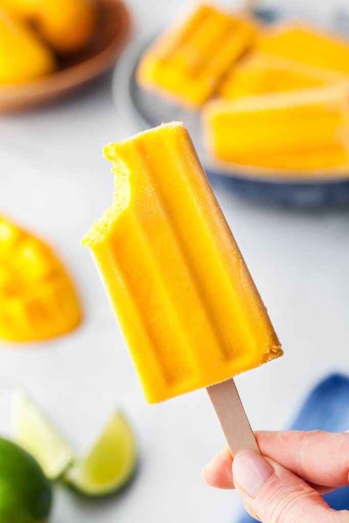 One mango popsicle with bite out of the top left corner being held up in the middle of the photo.  In the background there is a shallow bowl filled with ice containing other mango popsicles, and there is a sliced fresh mango on the left side in the background.