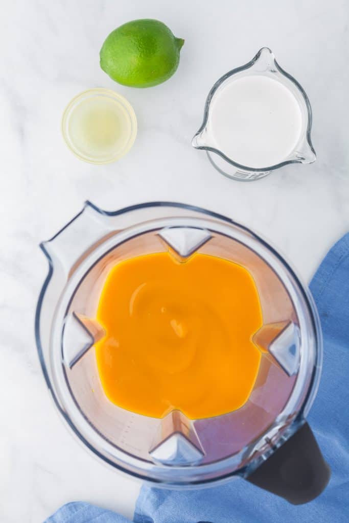 Blender filled with mango pulp in the centre.  Upper left corner is the lime juice in a small dish, and the right corner is a measuring cup filled with coconut milk
