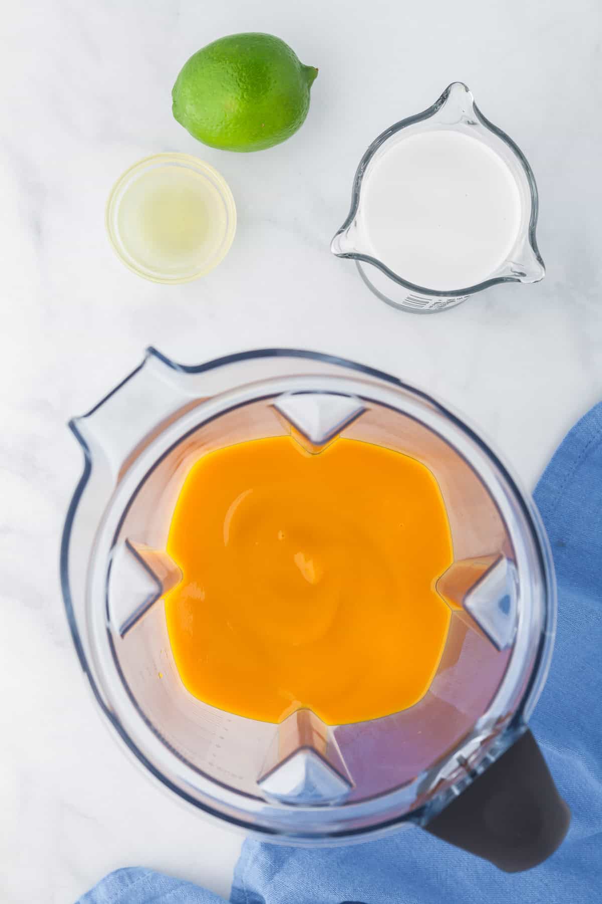 Blender filled with mango pulp in the centre. Upper left corner is the lime juice in a small dish, and the right corner is a measuring cup filled with coconut milk