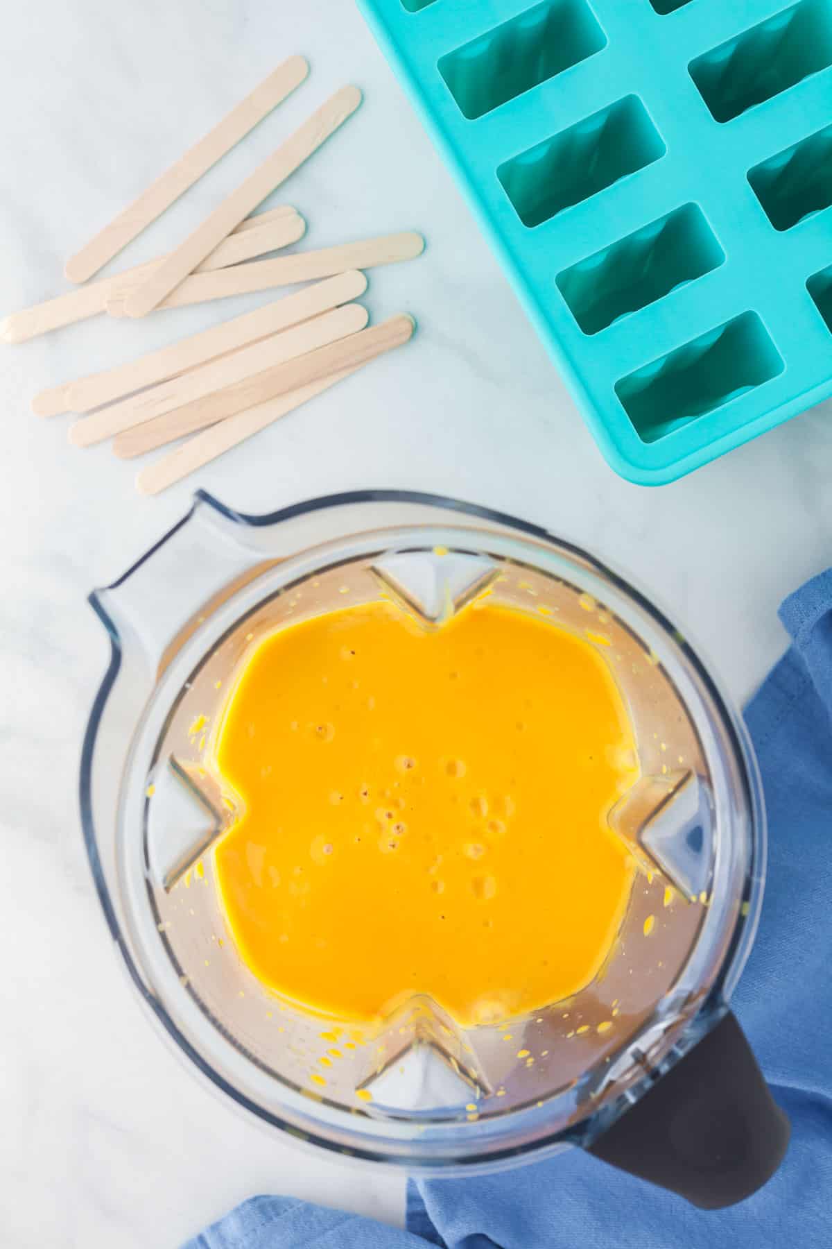 Middle of photo contains the blender filled with the homogenously mixed mango popsicle mixture. Upper left corner contains popsicle sticks and upper right of photo contains the silicone popsicle mold.