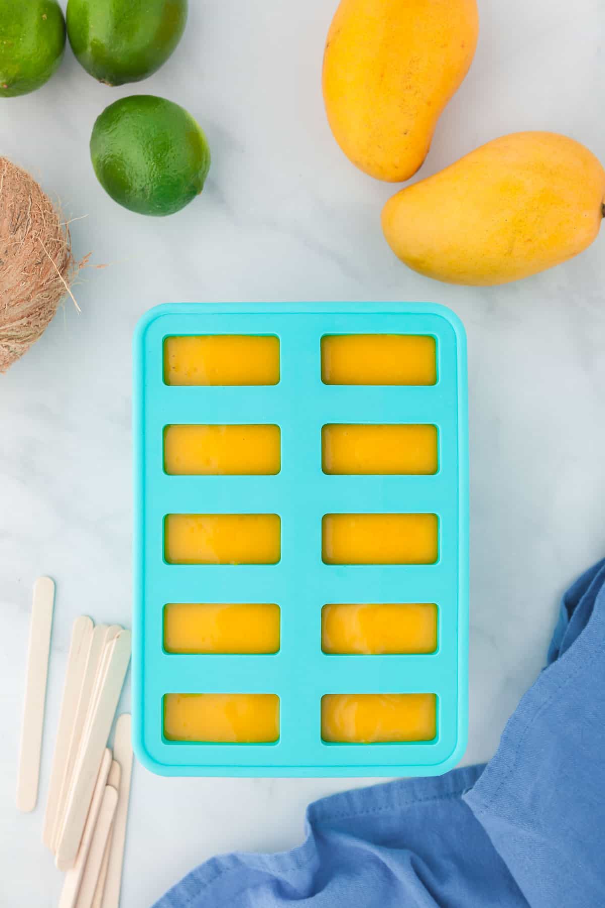 Popsicle mold has been filled with the mango popsicle mixture. Popsicle mold is surrounded by fresh mangoes, limes, coconuts, popsicles sticks and a blue linen.