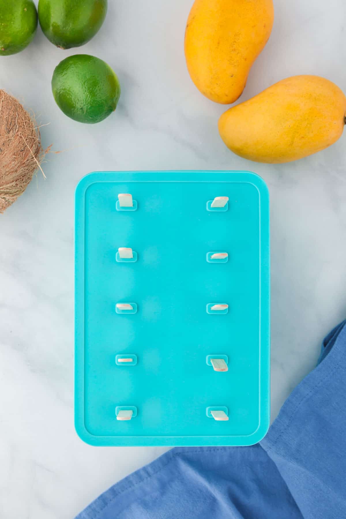Popsicle mold filled and lid placed on. Ten popsicle sticks inserted into the holes of the popsicle mold. Entire mold is surrounded by fresh mangoes, limes, and coconuts.