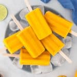 Overhead picture of a large blue bowl filled with ice and 6 mango popsicles laying on top. Top left corner has sliced limes, top right corner a blue linen, bottom left corner some coconut is peeking through, and the bottom right corner has fresh mangoes
