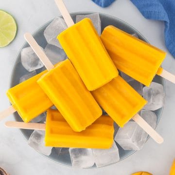 Overhead picture of a large blue bowl filled with ice and 6 mango popsicles laying on top. Top left corner has sliced limes, top right corner a blue linen, bottom left corner some coconut is peeking through, and the bottom right corner has fresh mangoes