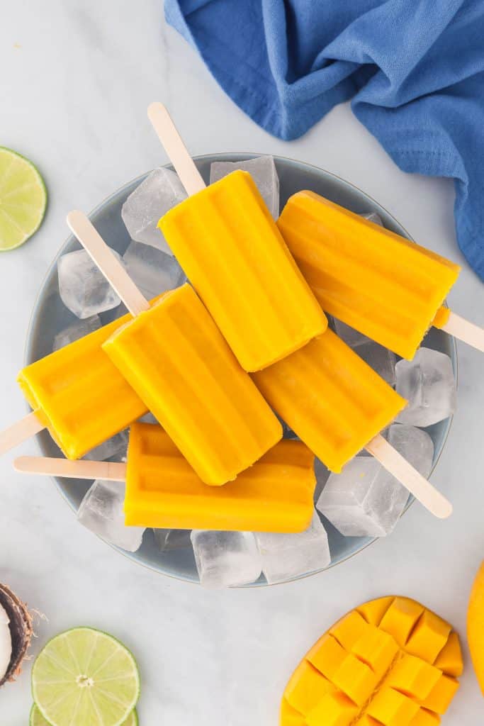 Overhead picture of a large blue bowl filled with ice and 6 mango popsicles laying on top. Top left corner has sliced limes, top right corner a blue linen, bottom left corner some coconut is peeking through, and the bottom right corner has fresh mangoes