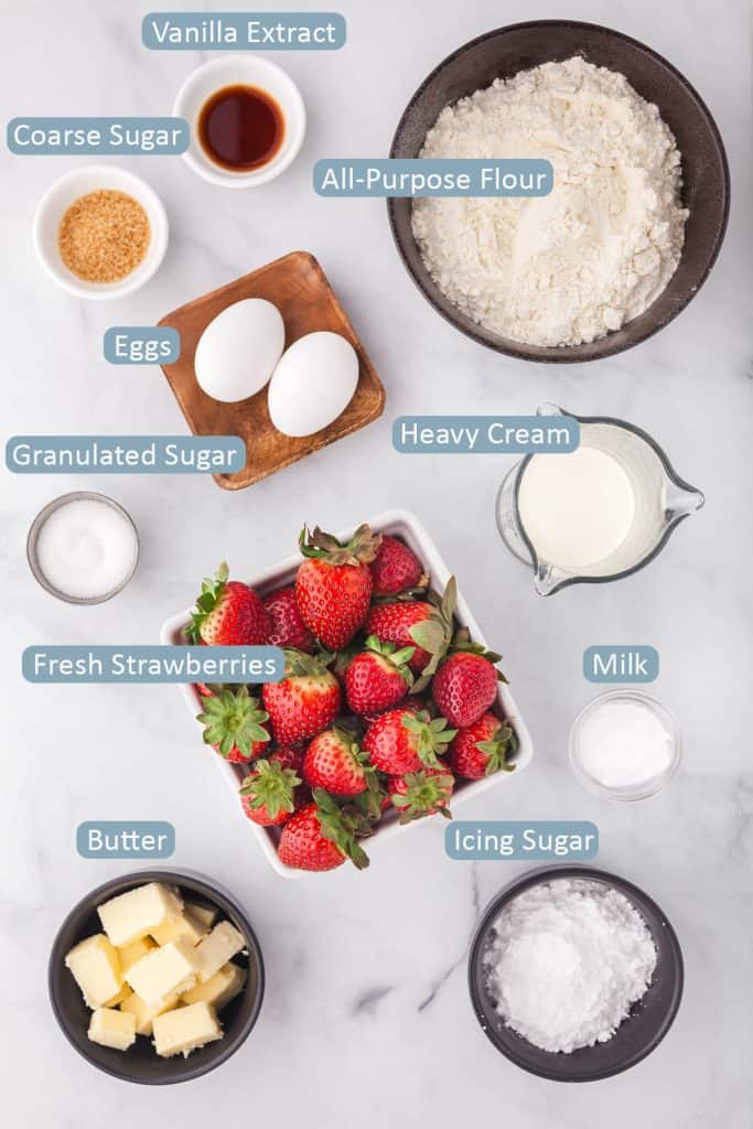 Overhead ingredient shot for strawberry scones including: fresh strawberries, all-purpose flour, vanilla extract, granulated and coarse sugar, eggs, heavy cream, milk icing sugar, butter.
