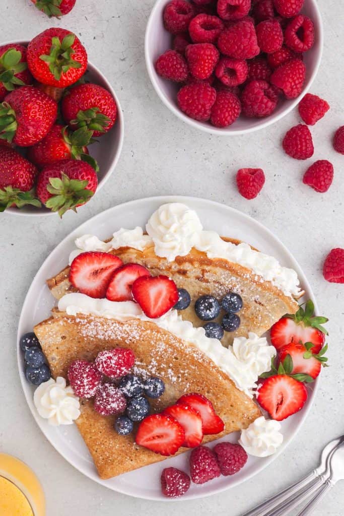 Overhead shot of a plate with two folded crepes on it, lightly filled with whipped cream and topped with raspberries, blueberries, and sliced strawberries. Top left corner has a small bowl filled with whole strawberries and the upper right corner has a small bowl of raspberries.