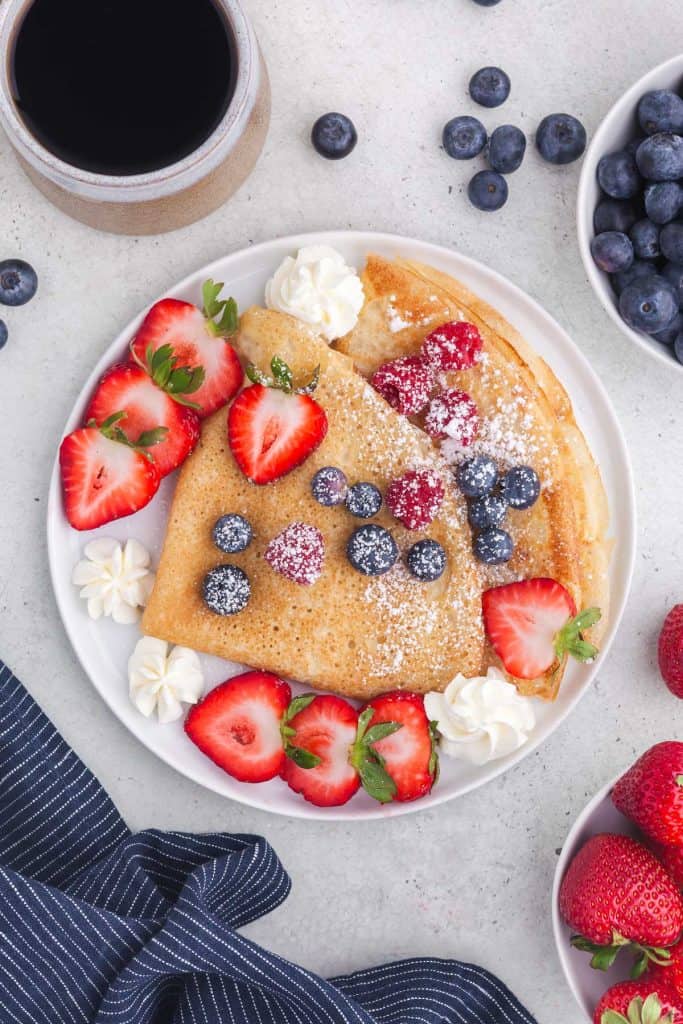 Two crepes folded into triangles and served on a white plate with sliced strawberries, whole raspberries and blueberries and sprinkled with icing sugar. A few dollops of whip cream on teh side.