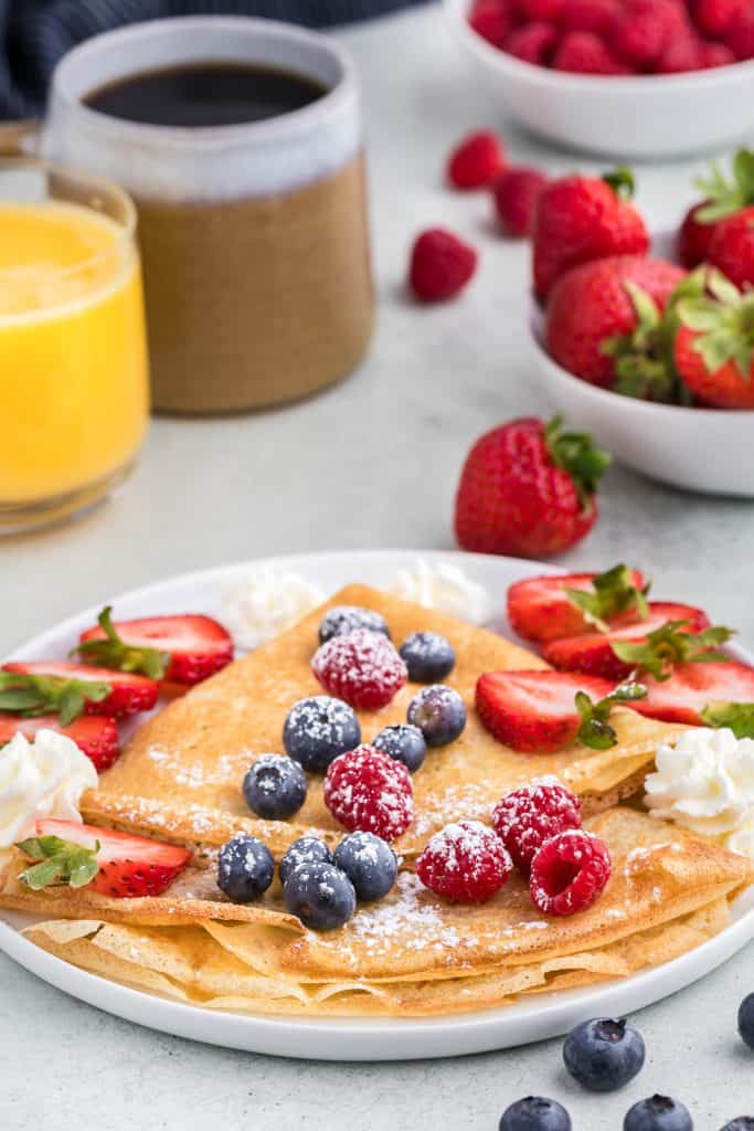 45 degree angle shot of plate filled with two folded crepes covered in raspberries, blueberries, and slice strawberries, as well as a few dollops of whipped cream and a sprinkling of icing sugar. Background in the upper left corner is a cup of orange juice and coffee and the upper right side is a small bowl filled with strawberries.