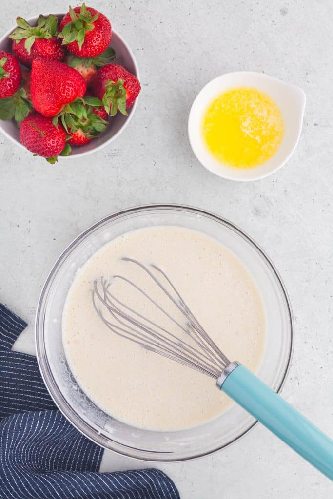 Overhead shot of crepe batter with a whisk in the bowl. Upper left corner is a bowl of strawberries and the bowl on the upper right is a small bowl of melted butter.
