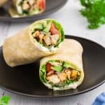 side view of a sliced chicken wrap. One half is laying on a black plate with it's open face facing the camera while the other half is propped up over the bottom half.