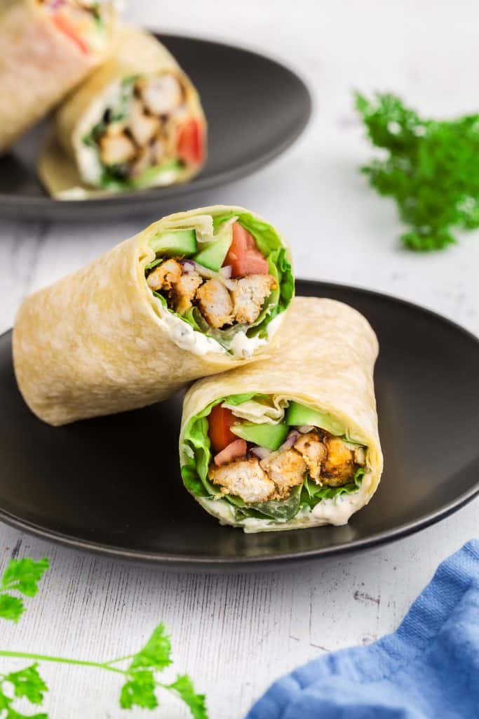 side view of a sliced chicken wrap. One half is laying on a black plate with it's open face facing the camera while the other half is propped up over the bottom half.