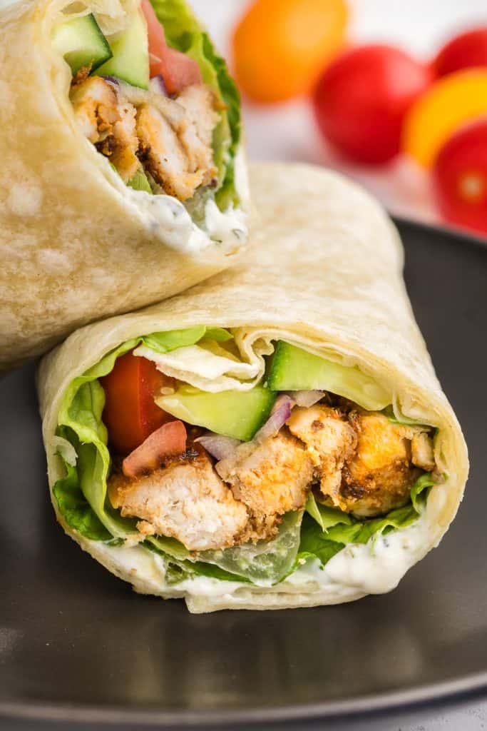Head-on photo of a chicken wrap sliced in half on a small black round plate. One half of the wrap has it's open face facing towards the camera and the other half is leaning on it at a 90 degree angle. You can see the chicken, lettuce, tomatoes, cucumber and ranch dressing inside.