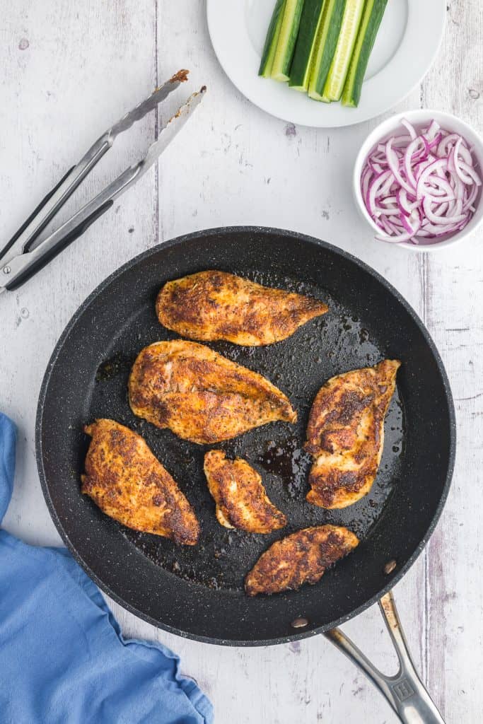 Overhead shot of seasoned chicken cooking in skillet for the chicken wraps. Upper left corner has a pair of tongs ready to flip the chicken and the upper right corner has a small white bowl of sliced red onions.