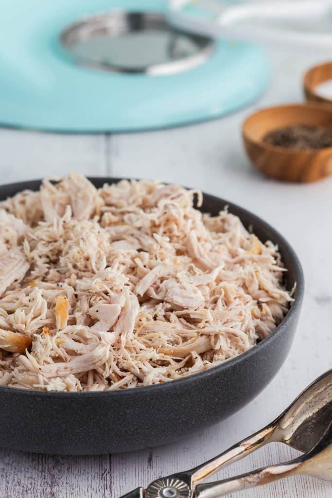 Side angle shot of a large bowl of dutch oven shredded chicken with the bottom of a stand mixer and salt and pepper wooden pinch bowls in the background.