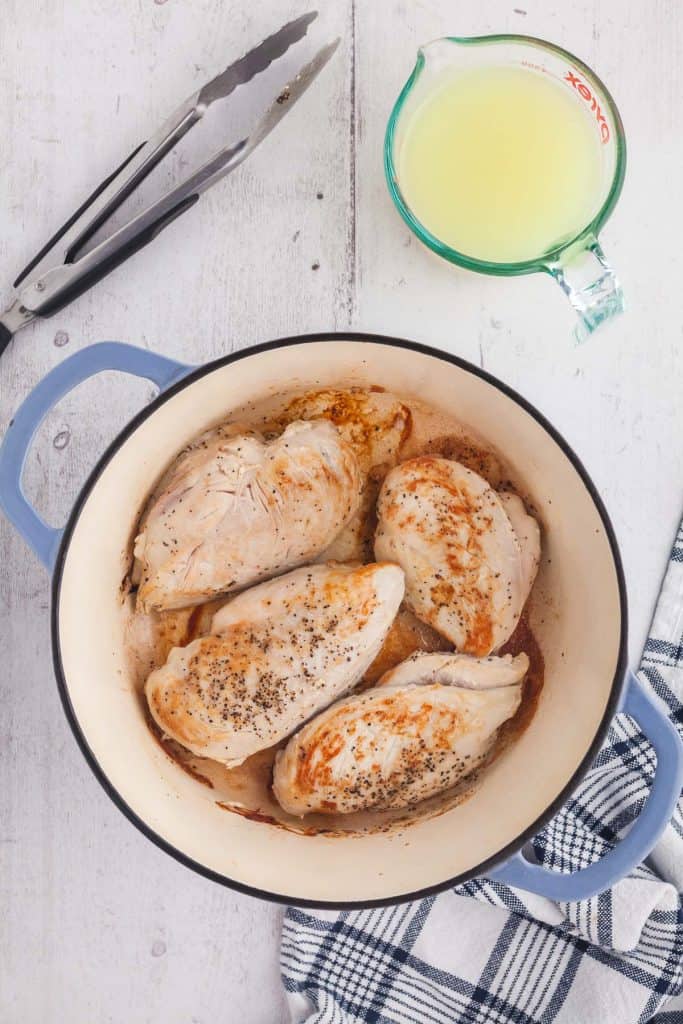 Overhead shot of chicken breasts searing in a dutch oven. Tongs for flipping the chicken are in the upper left corner and chicken broth the next ingredient is in the upper right corner