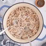 Overhead shot of shredded chicken in a dutch oven. Salt and pepper in wooden pinch bowls in the upper right corner, and a mixer paddle in the upper left corner