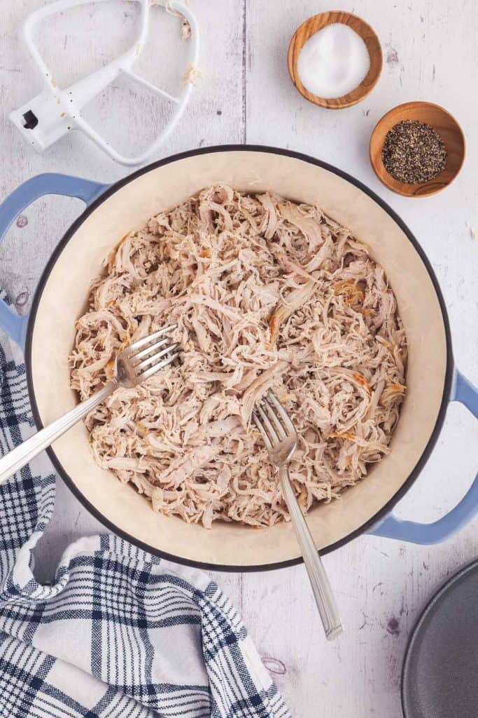 Overhead shot of shredded chicken in a dutch oven. Salt and pepper in wooden pinch bowls in the upper right corner, and a mixer paddle in the upper left corner
