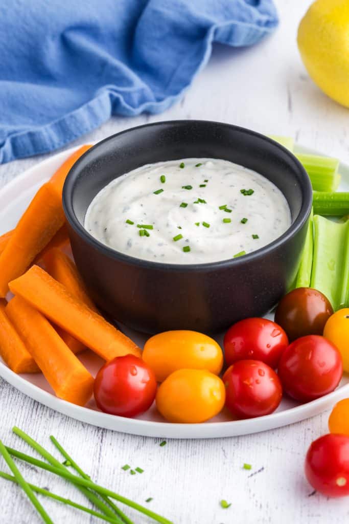 45 degree angle shot of a plate of veggies with a small black bowl in the middle filled with a creamy Greek yogurt ranch dressing for a dip. The top right corner has a lemon peeking in, the bottom right corner has two small cherry tomatoes peeking in to the scene and the bottom left corner has some peeking chives