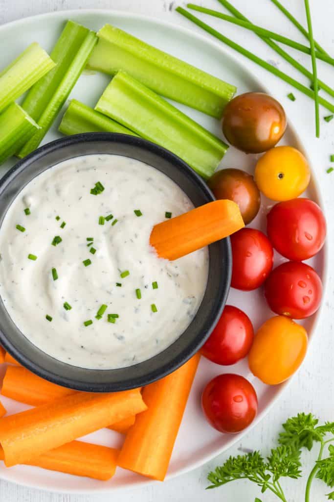Overhead shot of Greek yogurt ranch dressing in a small black bowl served on a white plate filled with cucumber, carrots, and cherry tomatoes.